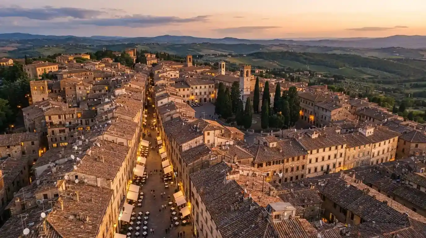 Vista dall'alto di una città italiana al tramonto, con luci che si accendono nelle finestre degli edifici lungo una via trafficata.